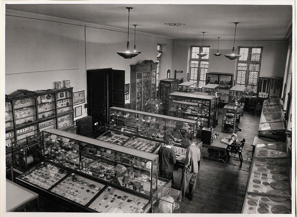 the top floor of the building, showing wooden exhibition cases of various sizes filled with objects. A woman is sitting at a desk reading, and a man and a woman are leaning over one of the cases.]