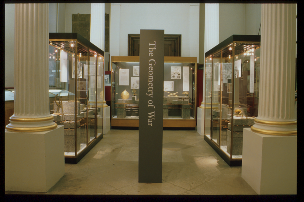 a photograph of the exhibition Geometry of War. A large pillar stands in the middle with the exhibition title on it. To either side, and behind pillars, are the first two exhibition cases
